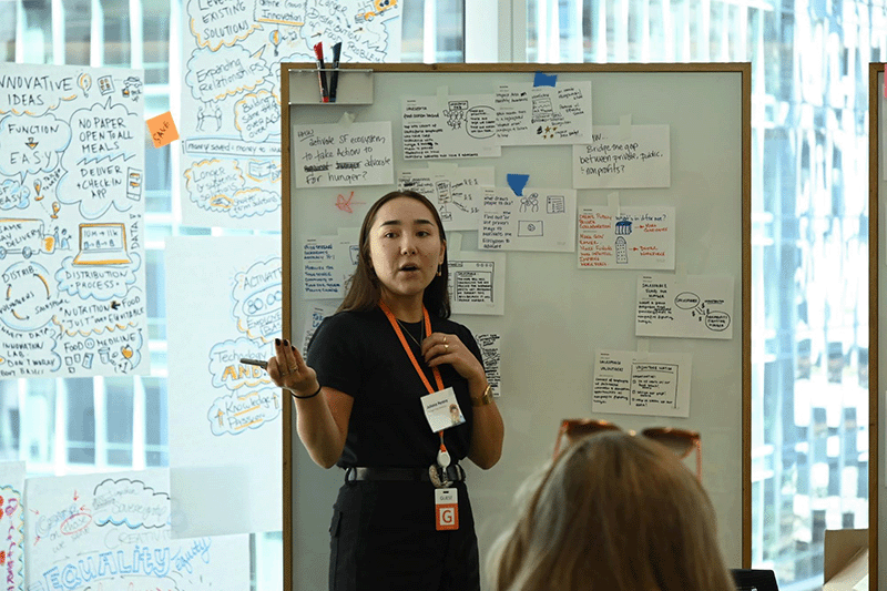 Women with a badge standing in front of a whiteboard brainstorming with an audience.