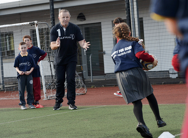 Sean Fitzpatrick teaching several children to play rugby  as a child runs holding a rugby ball