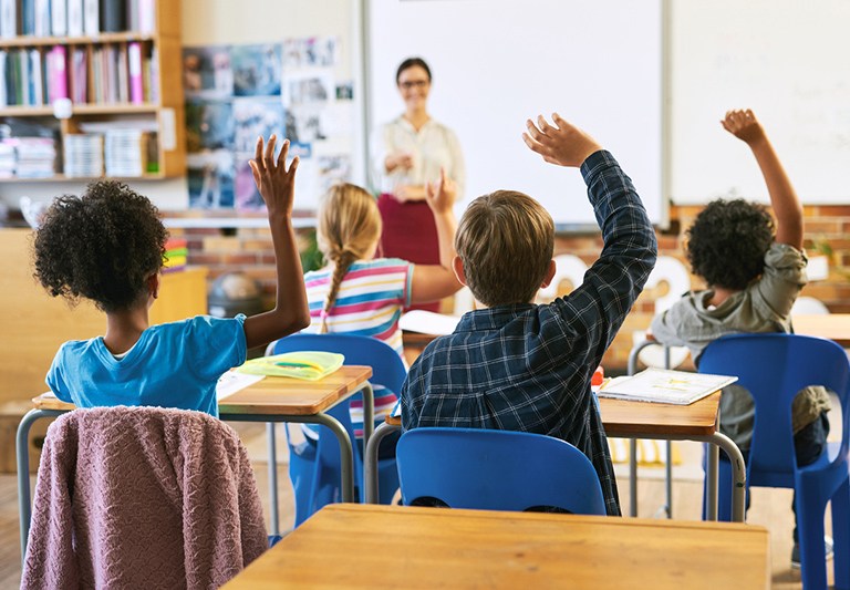 4 children sitting in a classroom raising their hands